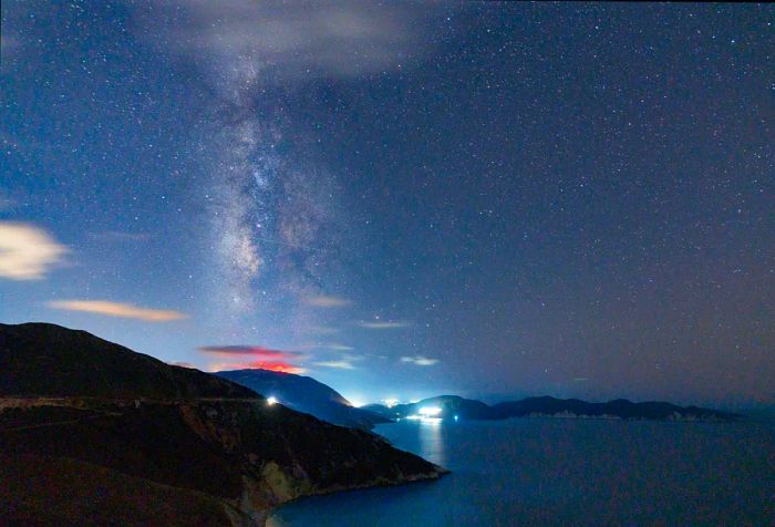 A car drives along a coastal road under the shimmering Milky Way in Greece.