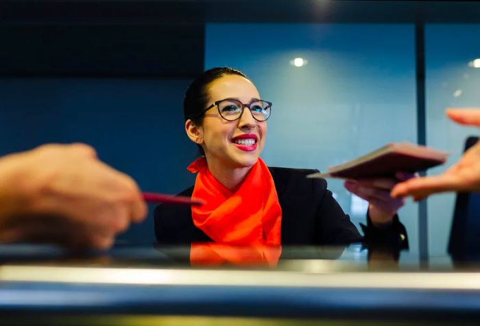 A friendly receptionist hands a passport to a traveler while another submits theirs at the counter.