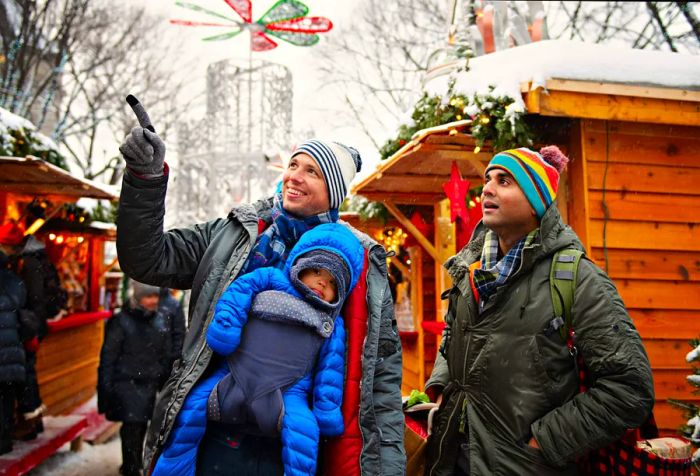 A couple with their child in a baby carrier, bundled up in winter attire, enjoying a snowy Christmas market.