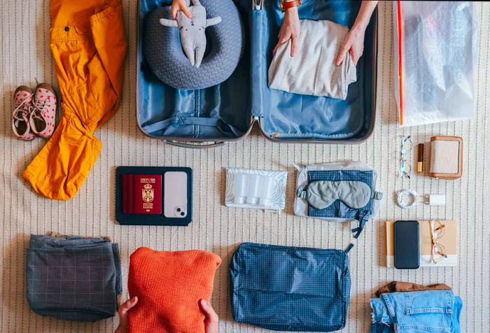 A flat lay showcasing a family preparing for their upcoming vacation, with an unidentifiable young girl placing a toy into a suitcase. Items like a passport, mobile phone, travel pillow, and more are arranged on the carpet.