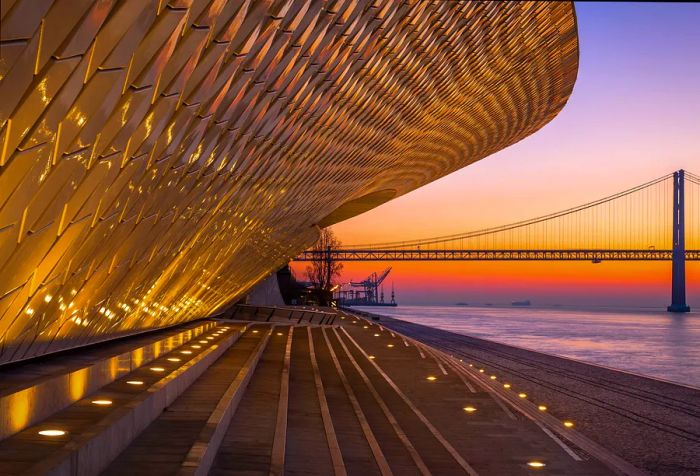 A uniquely designed museum illuminated along the riverbank, offering a stunning view of a long suspension bridge at sunset.