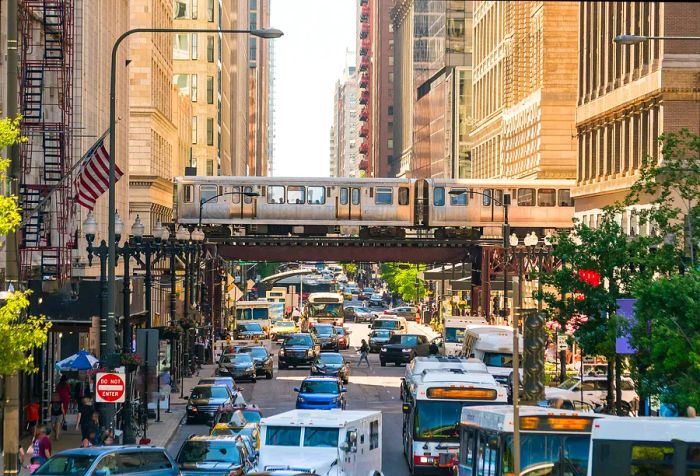 A gray train glides along an elevated track, passing over a bustling street amidst towering buildings.