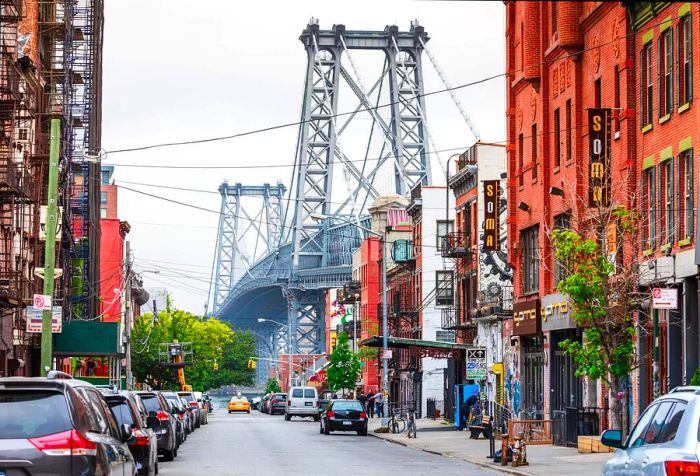 The two towering steel structures of a suspension bridge align prominently with a row of commercial buildings along the street.