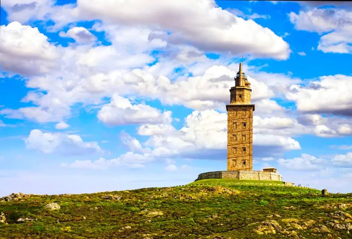 The Tower of Hercules stands as a tall lighthouse, featuring a spiral ramp outline atop a grassy hill.