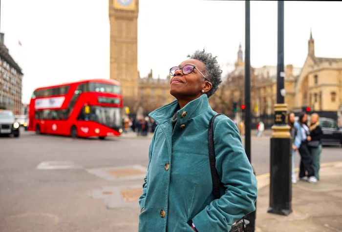 An elderly Black female tourist gazes up at the architecture in London, exploring the City of Westminster alone on a dreary winter day. Behind her stands the iconic Big Ben as she admires the view.