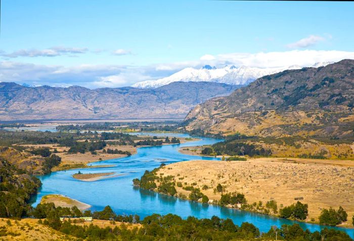 A sweeping view of a riverbed surrounded by lush greenery in a mountainous valley, with a snow-capped peak in the distance.