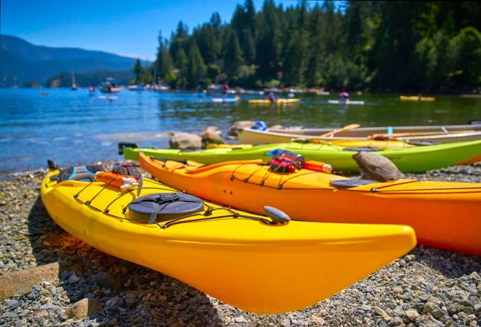 Yellow and green canoes are lined up along the pebbled shoreline of a bay, with people enjoying activities in the background.