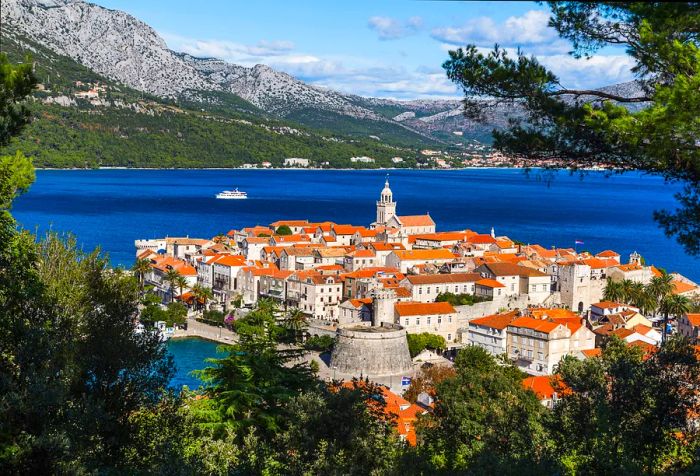 A charming ancient town featuring traditional stone houses and buildings that extend into the turquoise waters, framed by a verdant mountain range.