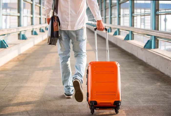 A man dressed casually, carrying a backpack and pulling a suitcase, strolls along a pathway leading to a train station.