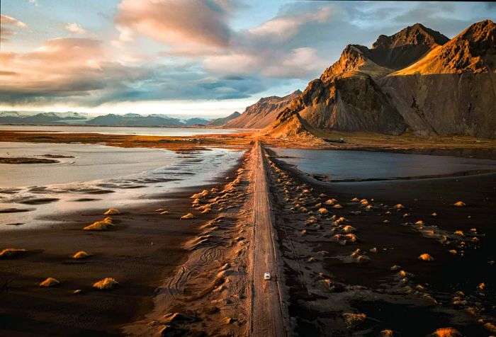 A white car navigates a rugged, narrow road through black sands, heading toward steep, pointed mountains.