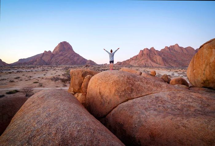 A woman stands triumphantly on a large rock in the mountains, arms raised high to the sky.