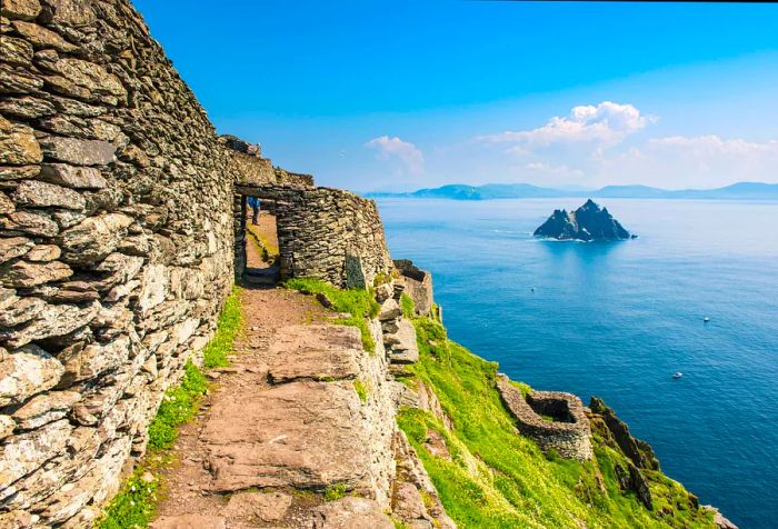 An ancient stone structure clings to a steep hillside, offering breathtaking views of the sea, while a striking rock formation rises from the water.