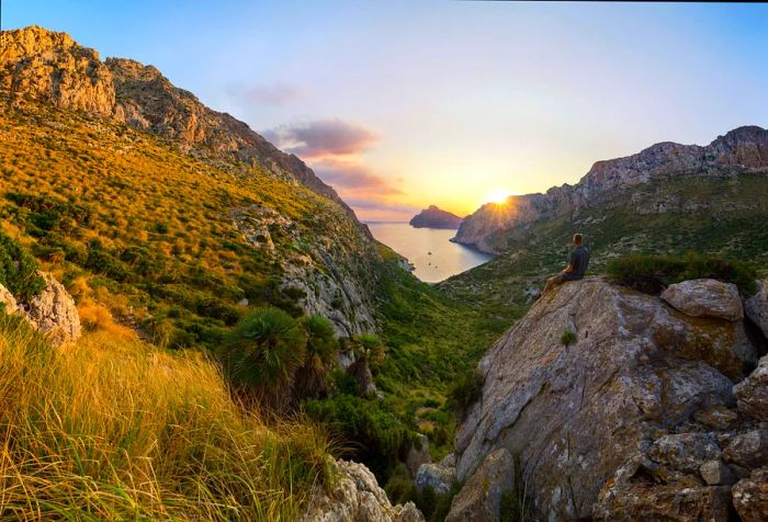 A man sits on a boulder, watching the sunrise through the mountains near a grassy slope.