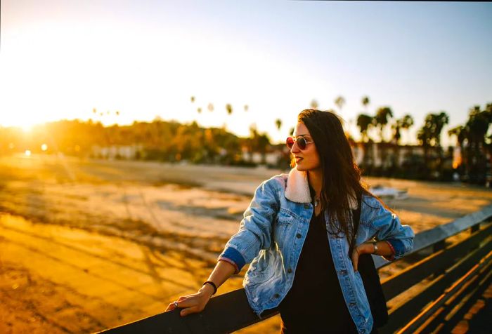 A long-haired woman in a denim jacket shields her eyes from the sun with sunglasses as she leans against a wooden fence on the boardwalk.