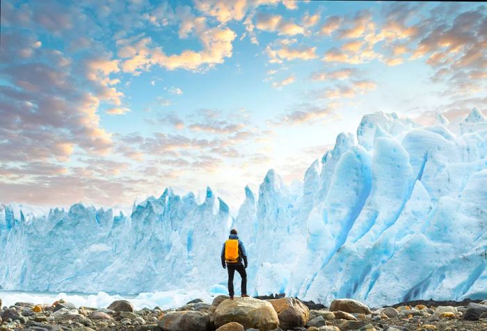 A hiker with a yellow backpack stands on a rock, gazing at towering ice formations.