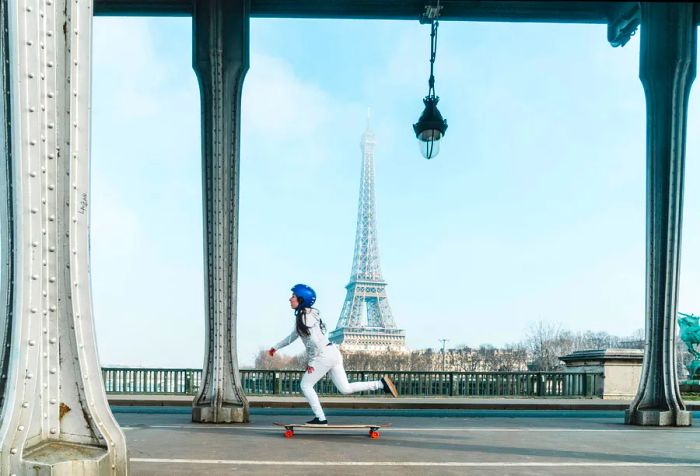 A skateboarder in a blue helmet glides down a street with the Eiffel Tower visible in the distance.