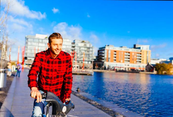 A young man in a red flannel shirt pedals his bike alongside a city canal, with buildings visible in the background.