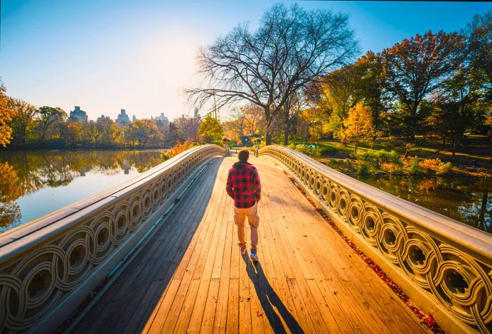 A man in a checkered shirt walks across a footbridge over a serene lake framed by colorful autumn leaves.