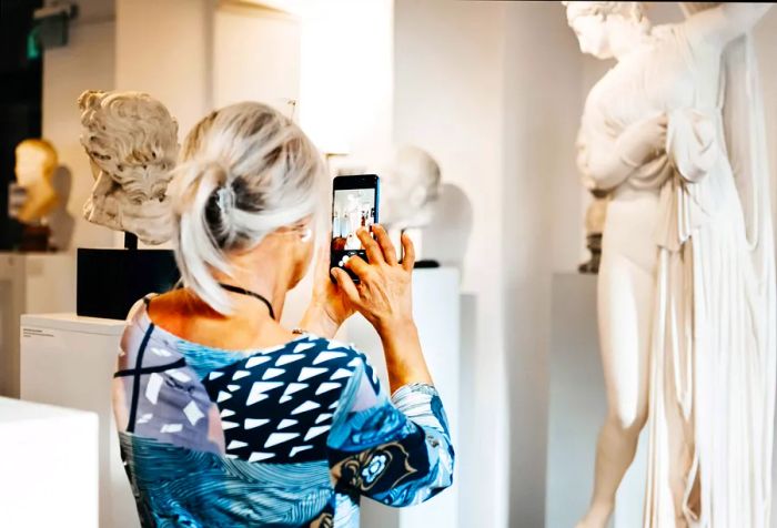 An older woman skillfully captures the essence of a classical sculpture using her smartphone.