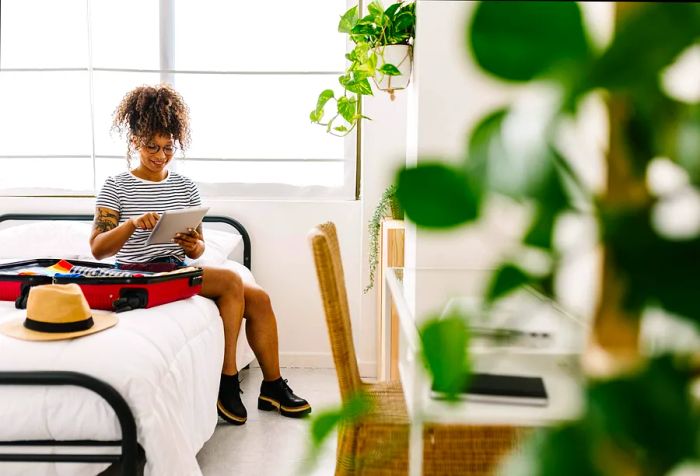 A woman sits on her bed with luggage, engaging with her tablet computer.