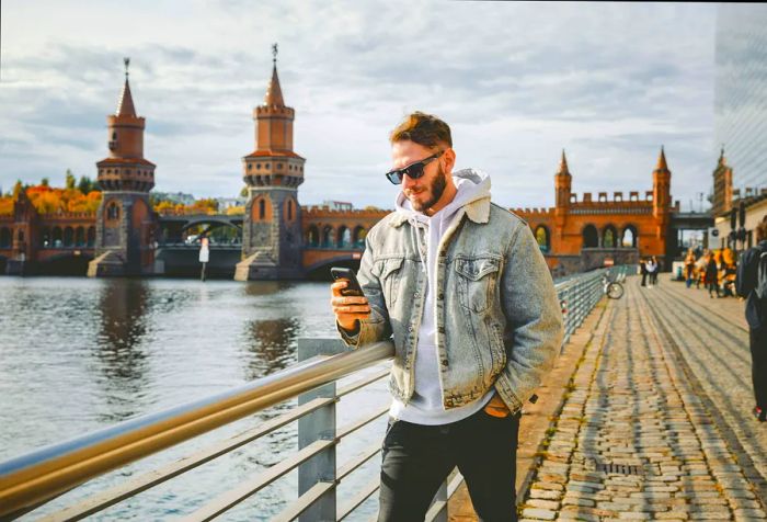 A man wearing sunglasses, a denim jacket, and black pants leans against a railing on a promenade while checking his smartphone.