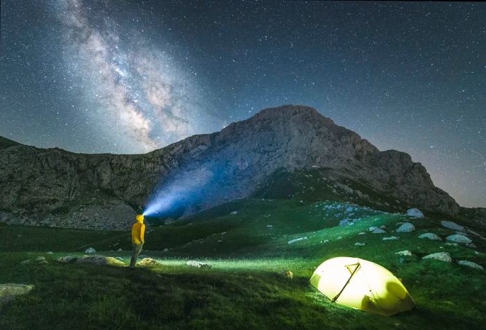 A nighttime scene featuring a man with a headlamp beside a glowing tent, set against a backdrop of twinkling stars.