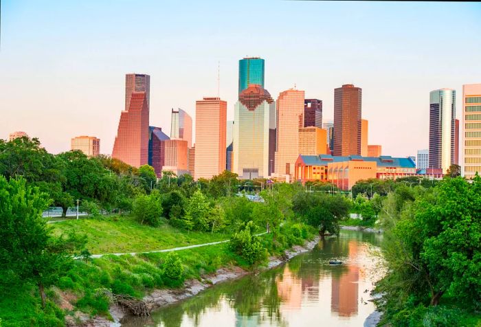 A serene stream, surrounded by vibrant greenery, flows toward a cluster of sleek skyscrapers beneath a bright sky.