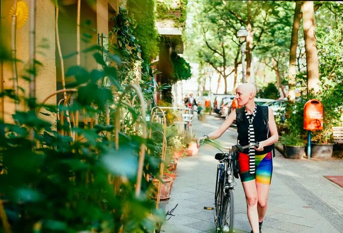 A bald person rides a bicycle along a street lined with buildings draped in lush greenery and tall trees.