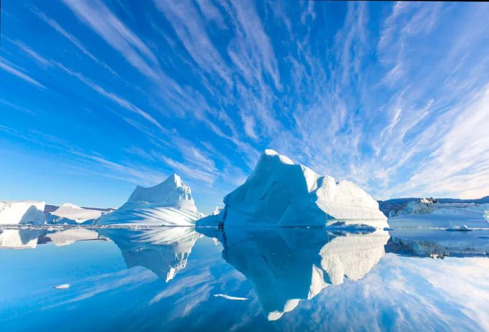 A serene view of icebergs mirrored in tranquil waters beneath a clear blue sky on a sunny day.