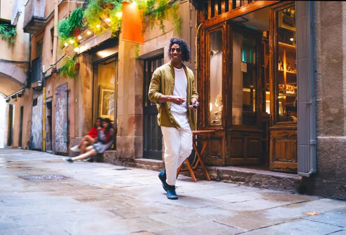 A man dressed in a white outfit and brown blazer strolls joyfully along a cobbled street, passing by classic building stalls.