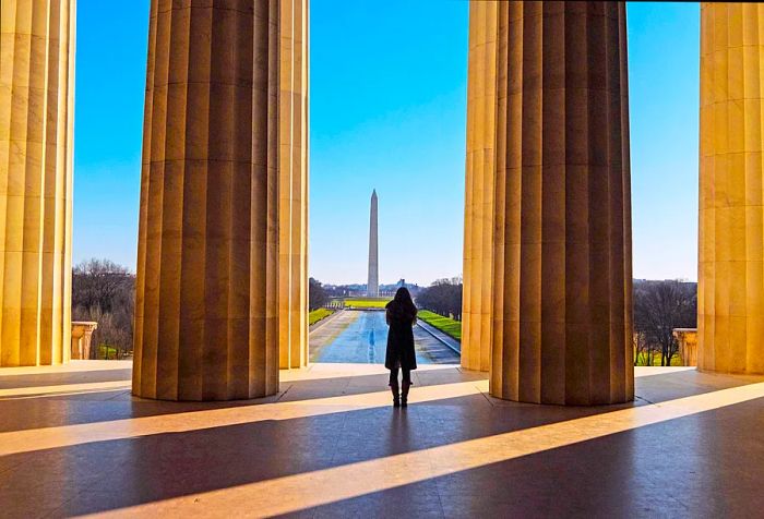 A woman stands between towering fluted columns, with the Washington Monument visible in the background.