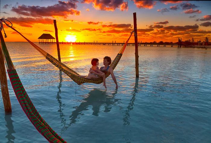 A picturesque scene of a mother and daughter relaxing in a hammock on the beach with a stunning sunset backdrop.