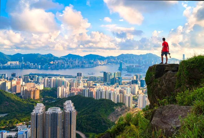 A male photographer stands at the edge of a mountaintop, gazing over the vast cityscape under a dramatic sky.