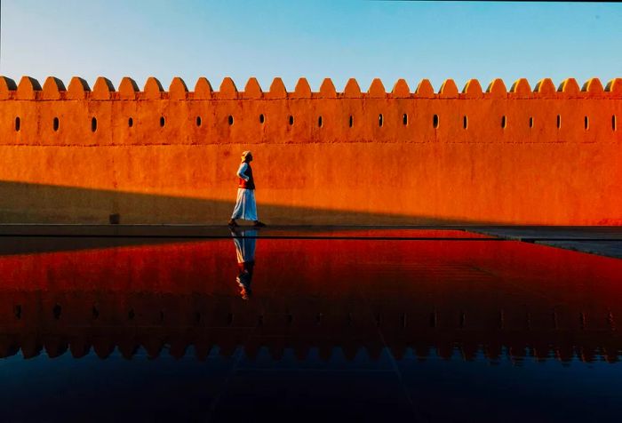 A man strolls along a fortress wall, his reflection visible in the tranquil pool of water beside him.