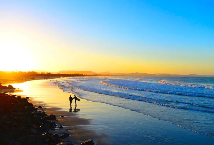 A silhouette of two surfers strolling along the beach, with gentle waves caressing the shore, framed by a stunning sunset.