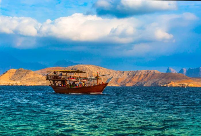 A dhow carrying passengers glides across the sea, with sandy mountains rising in the background.