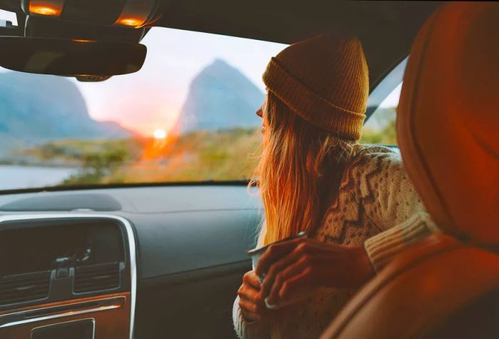 A woman with long hair wearing a cap sits in the passenger seat of a car, holding a cup of coffee.