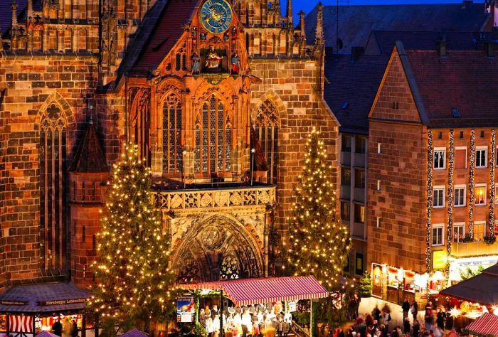 A stunningly adorned brick cathedral in Gothic architecture, flanked by towering Christmas trees at its entrance, with festive shops lining the main square.