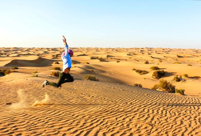 A male traveler leaps joyfully with arms raised in the heart of the desert.