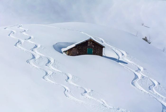 A cozy cabin nestled in deep snow, with visible ski tracks winding through the landscape.