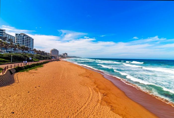 An unoccupied beach with crashing waves alongside a beautifully landscaped promenade and waterfront structures.