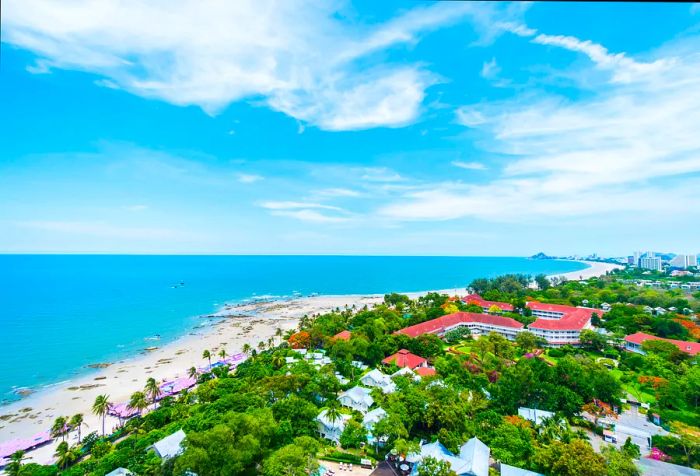 A charming coastal city featuring a white sandy beach next to the calm turquoise sea under a cloudy blue sky.