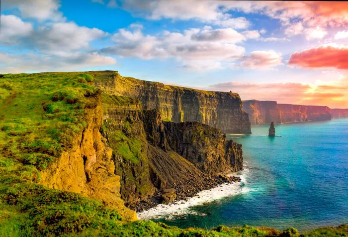 A woman and her dog stand on a grassy ledge atop the Cliffs of Moher.