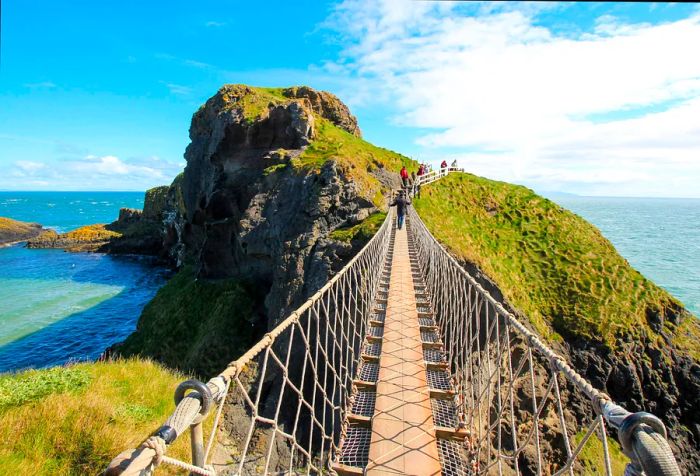 A slender, winding rope bridge connects to lush, rugged cliffs that tower over the ocean.