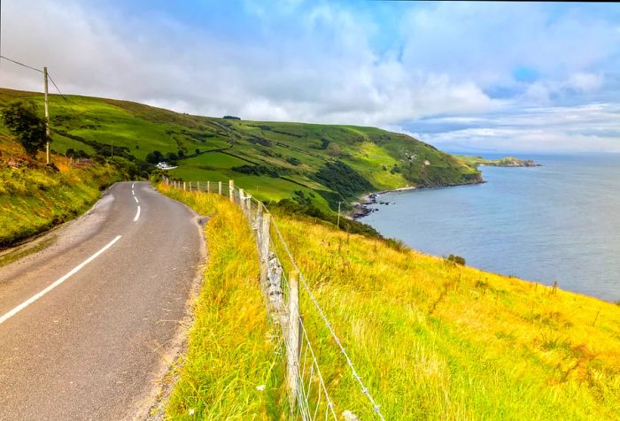 A coastal road winds along a hillside, bordered by a barbed-wire fence next to a grassy field.