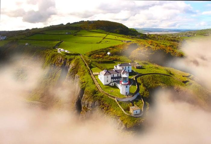 A lighthouse perched on a cliff's edge, surrounded by vibrant grasslands, with mist lingering along the hills.