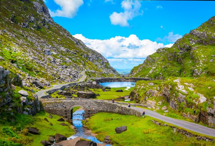 People traverse a stream on a stone bridge situated in a valley surrounded by rocky slopes.