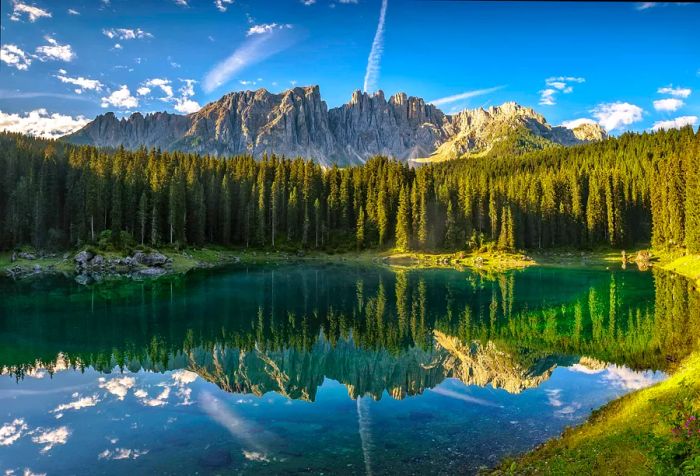 A stunning lake with crystal-clear waters reflects the surrounding trees and majestic mountains under a bright blue sky.