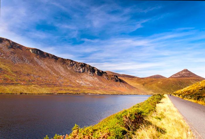 A paved pathway runs by the dam, nestled beneath the rugged mountain range.