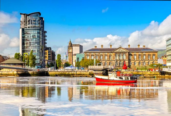 A fishing boat is anchored in a river, with historic harborfront buildings and a clock tower in the background.